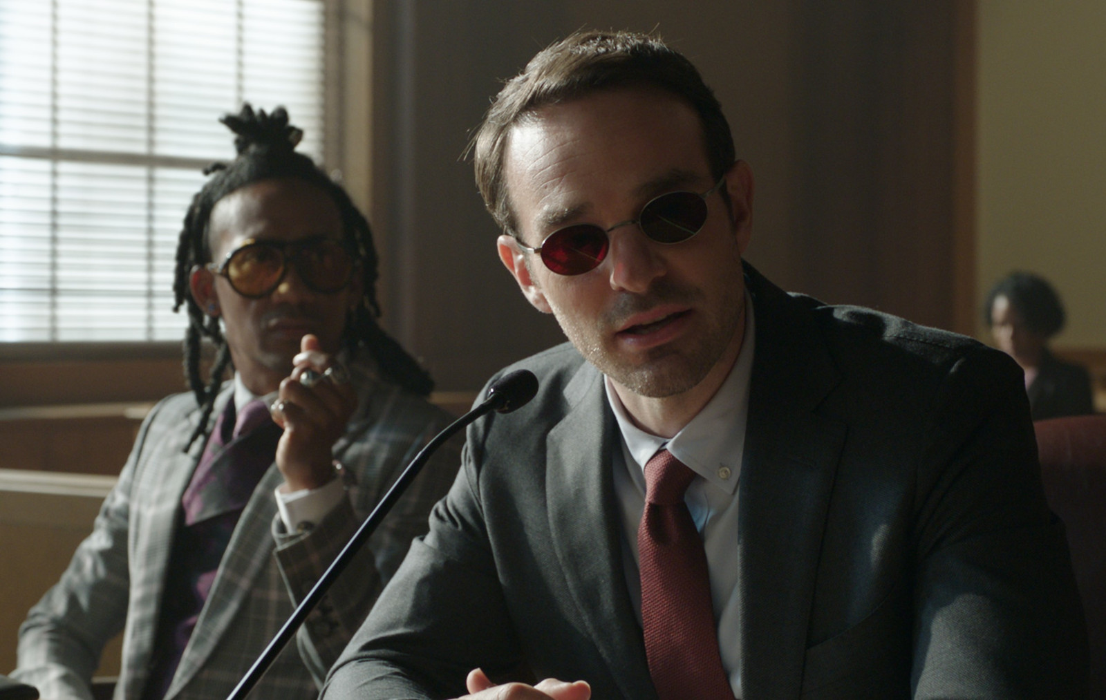 Griffin Matthews as Luke Jacobson and Charlie Cox as Daredevil/Matt Murdock in Marvel Studios' She-Hulk: Attorney at Law for our article about the 'Daredevil: Born Again' production schedule and premiere. They're sitting in the courtroom and staring at something off-screen.