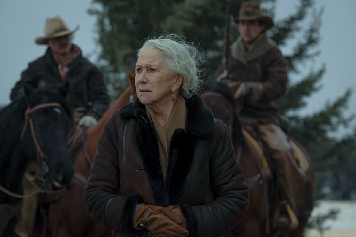 In 1923, Cara Dutton stands in front of two men on horses. She is wearing a coat and gloves.