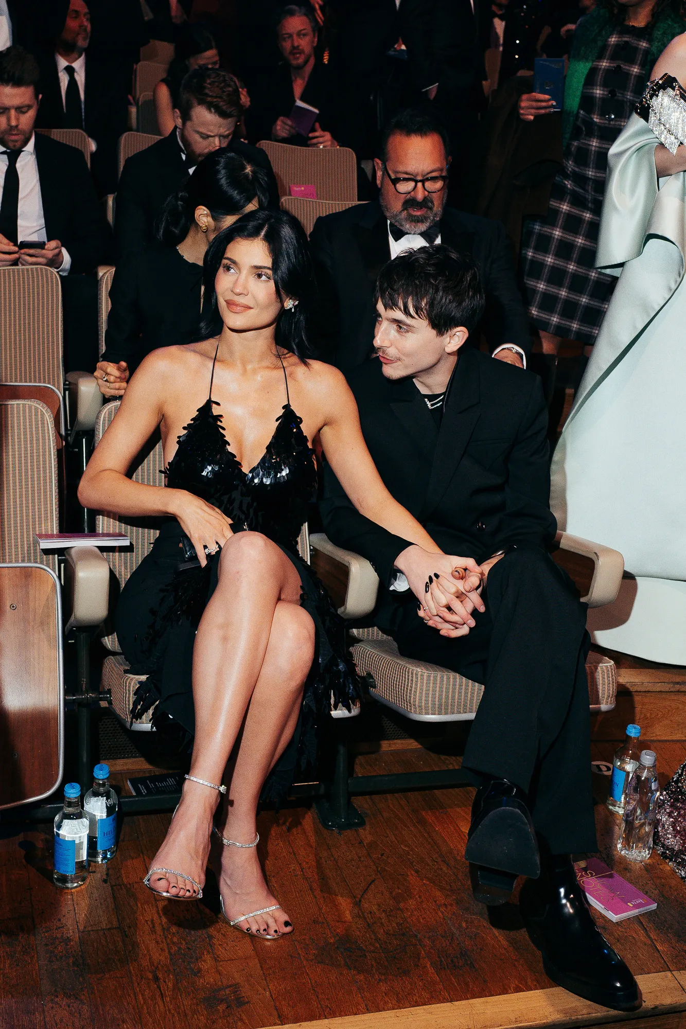 Kylie Jenner and Timothée Chalamet sitting together at the BAFTA Film Awards. Jenner is wearing a black dress and Chalamet is looking at her while holding her hand.