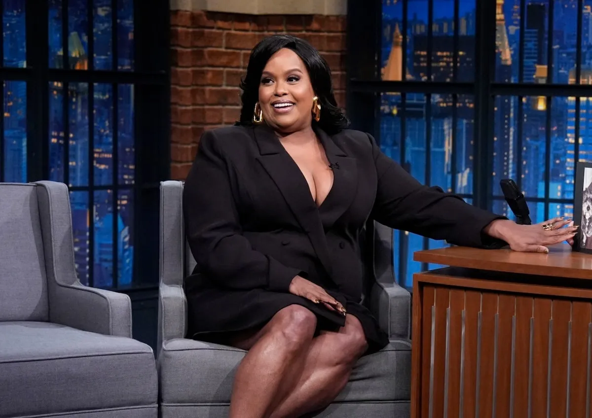 Wearing a black tuxedo dress, Natasha Rothwell smiles while Seth Meyers interviews her about The White Lotus