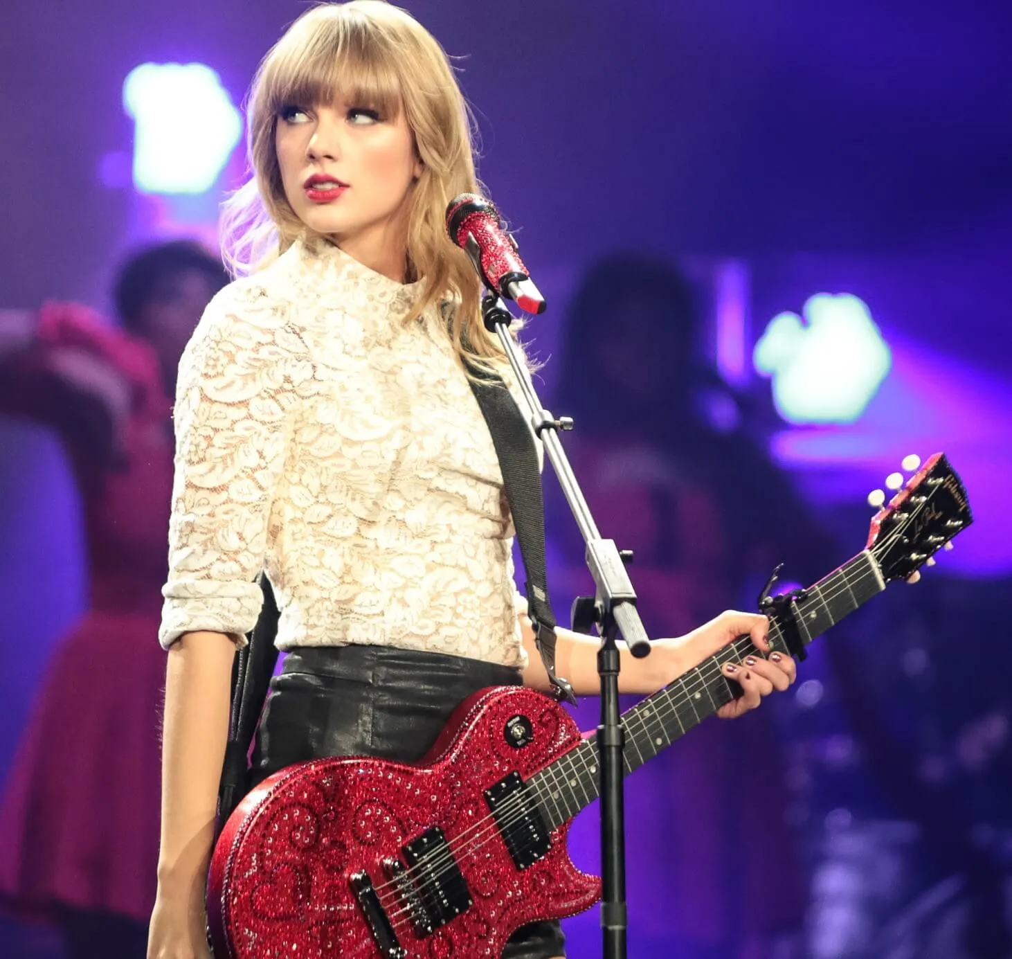 Taylor Swift stands on a stage with a red guitar. She wears a white shirt and stands in front of a microphone.